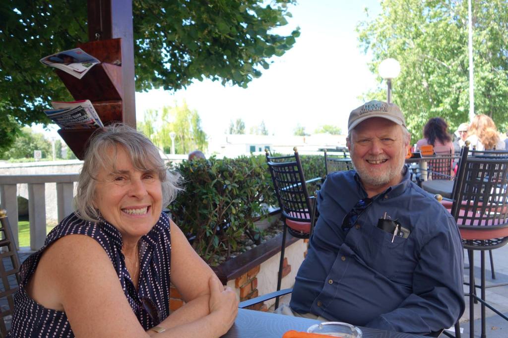 Jesse and Nancy are seated at an outdoor restaurant table in the shade. Both are looking to the camera and smiling.